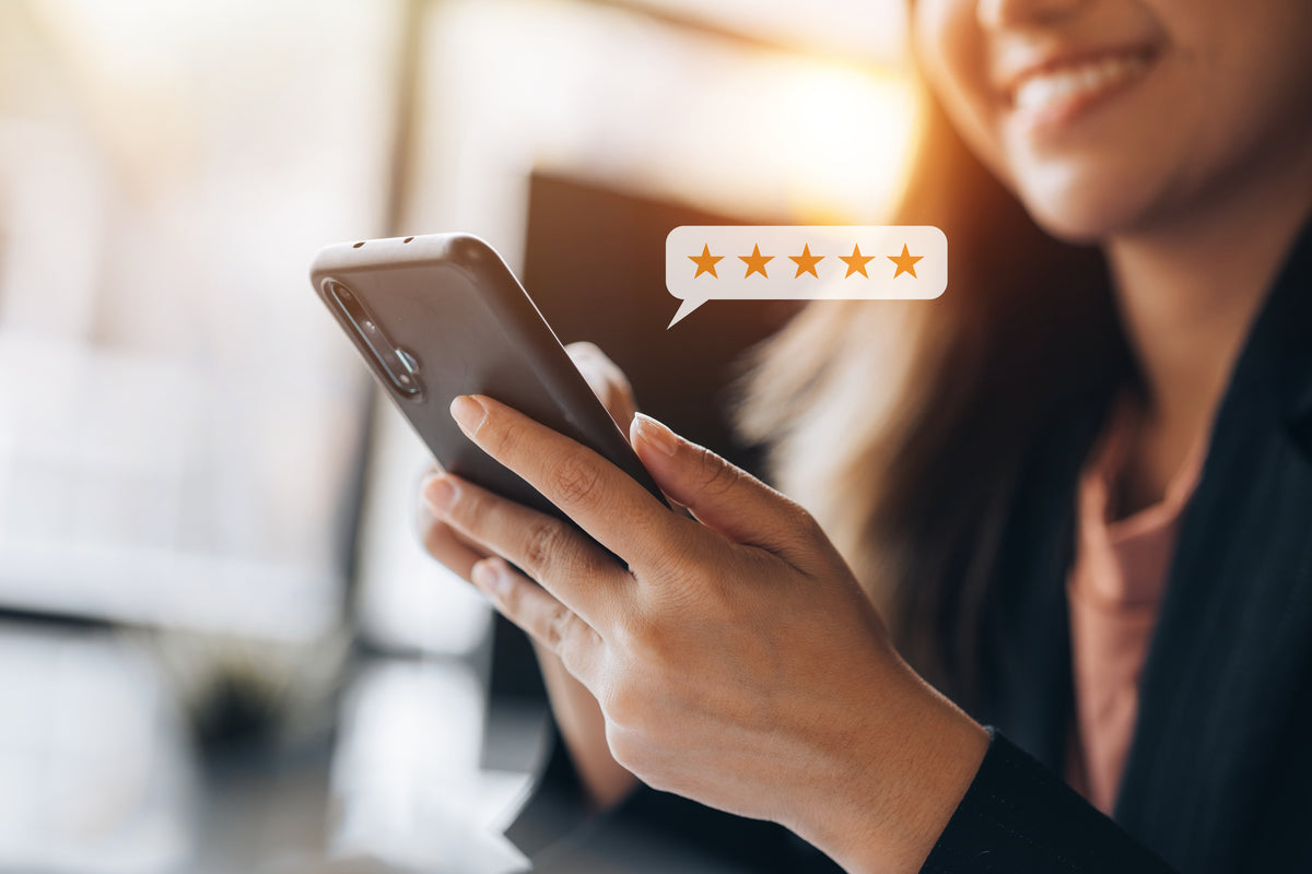 A close-up lifestyle shot of a woman holding a smartphone, with a speech bubble graphic showing five gold stars floating near the screen. The background is a warm, softly blurred indoor setting with bright sunlight.