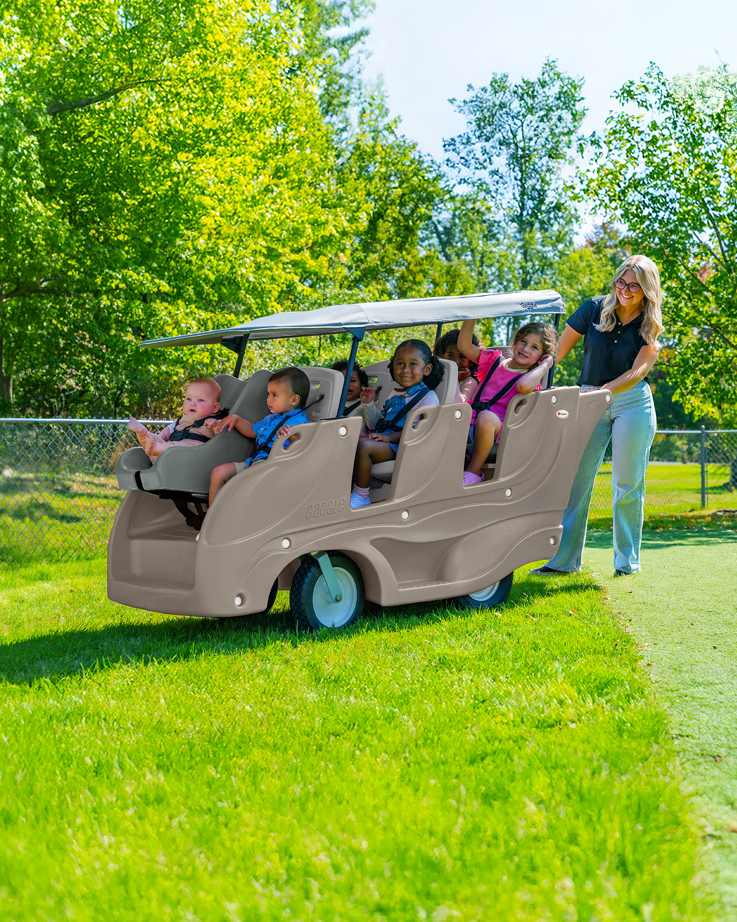 A woman pushes a beige four-seater golf cart-style ride-on toy outdoors on a grassy lawn, with children seated inside. Trees, grass, and a chain-link fence are visible in the background.