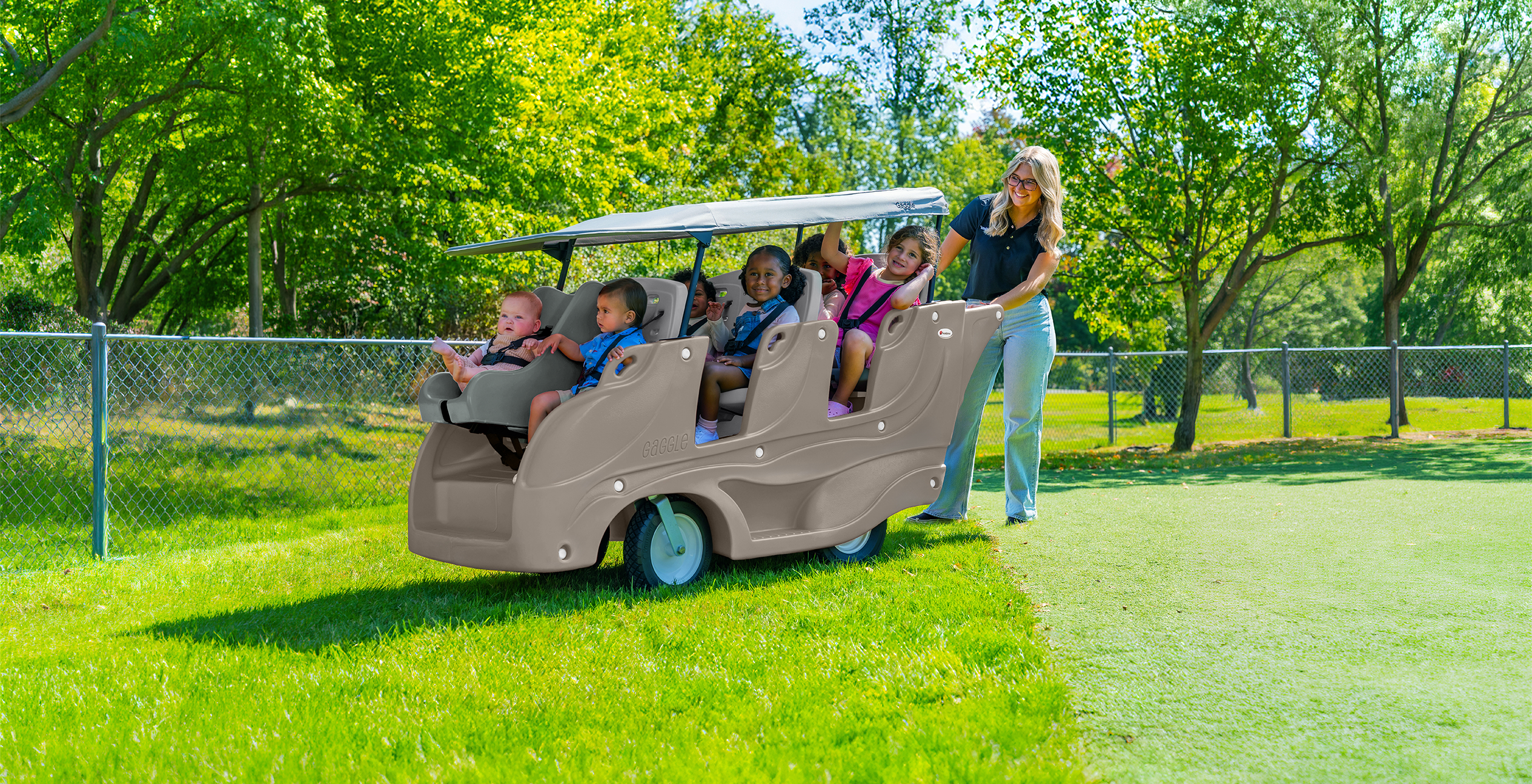 A woman pushes a beige four-seater golf cart-style ride-on toy outdoors on a grassy lawn, with children seated inside. Trees, grass, and a chain-link fence are visible in the background.