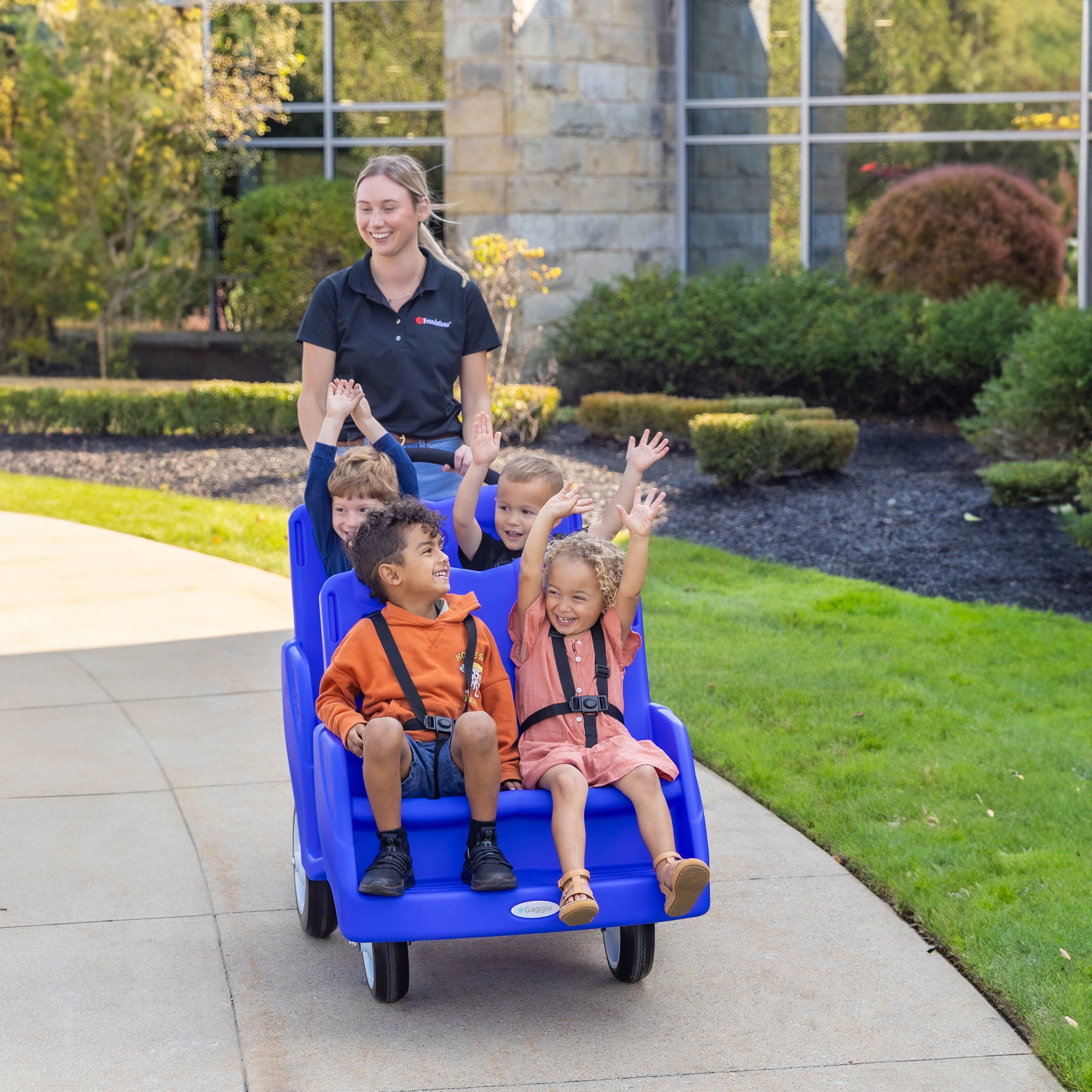 Children in a blue four seat buggy with a woman on a sidewalk, surrounded by greenery and a building.
