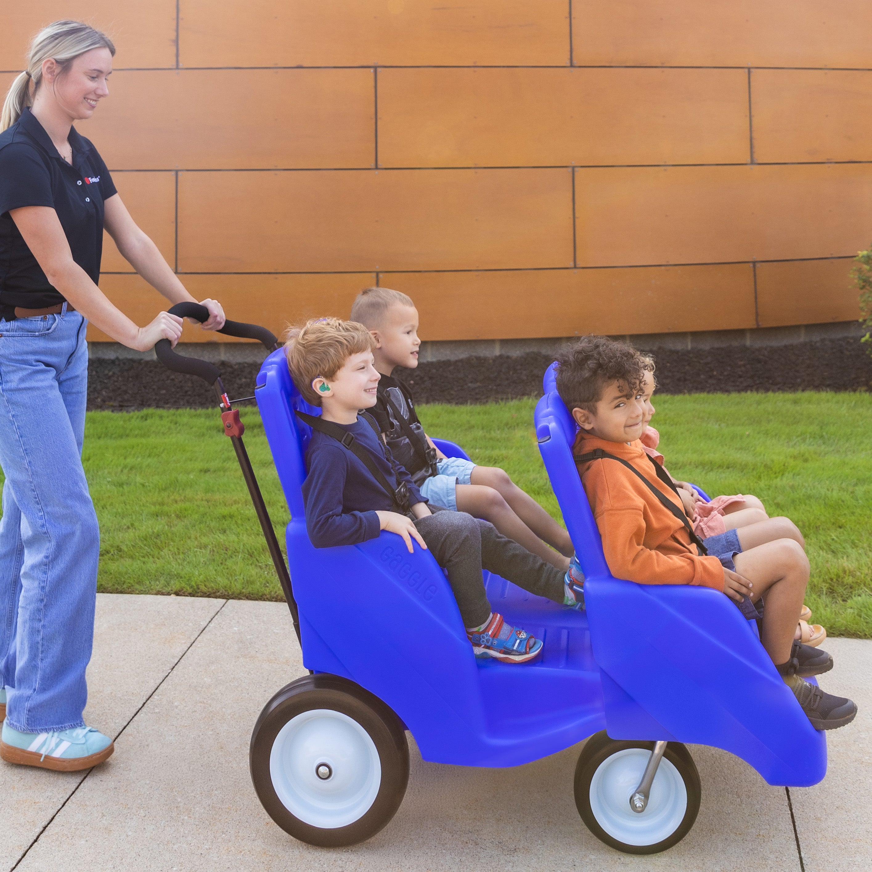 Woman pushing a blue buggy with four children sitting in it on a sidewalk.