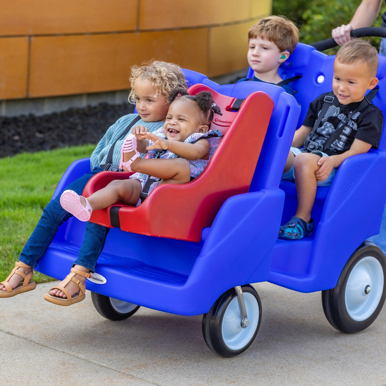 Children sitting in a blue 4 seat buggy with a red infant seat on a sidewalk.