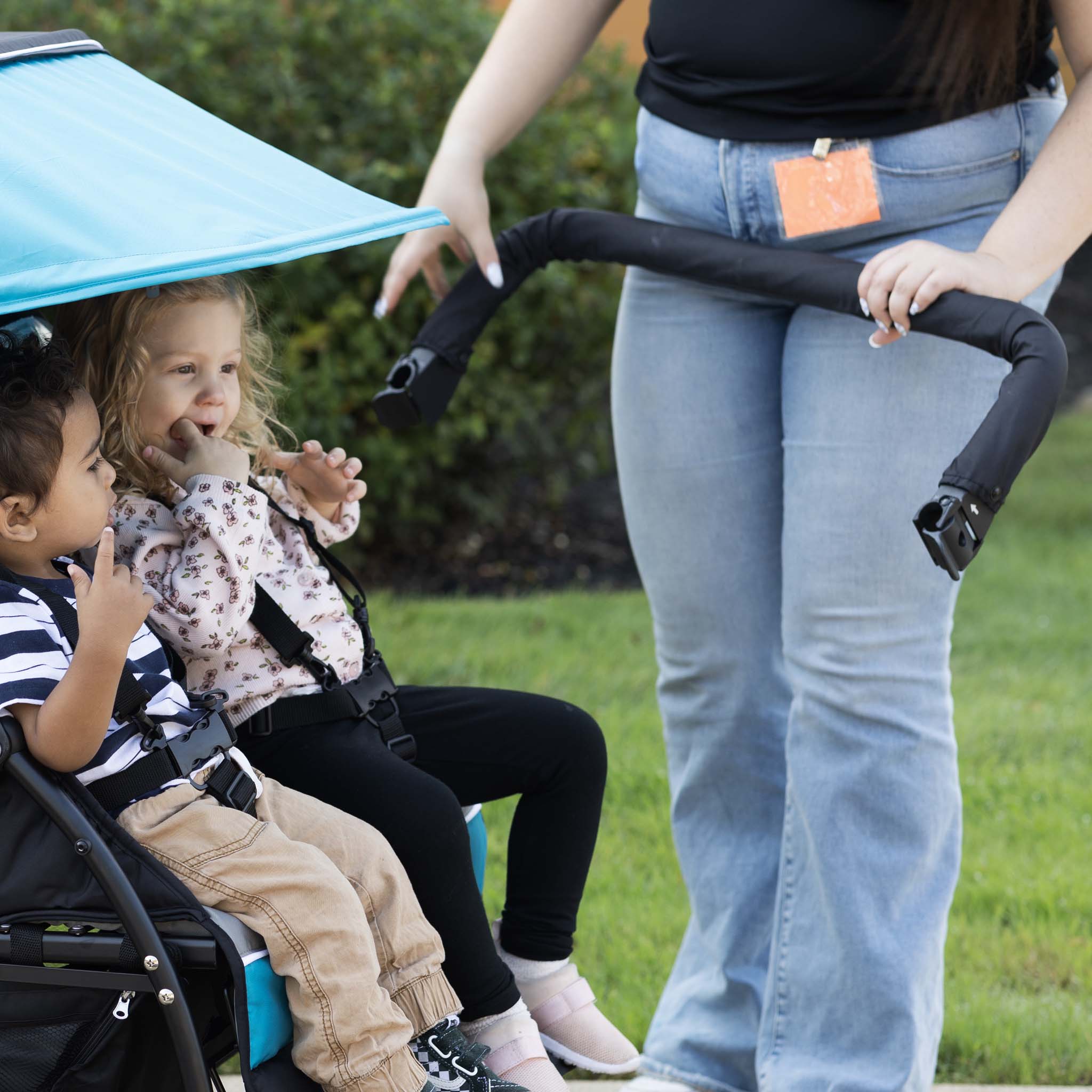 Two children in a stroller with a woman adjusting the arm bar outdoors.