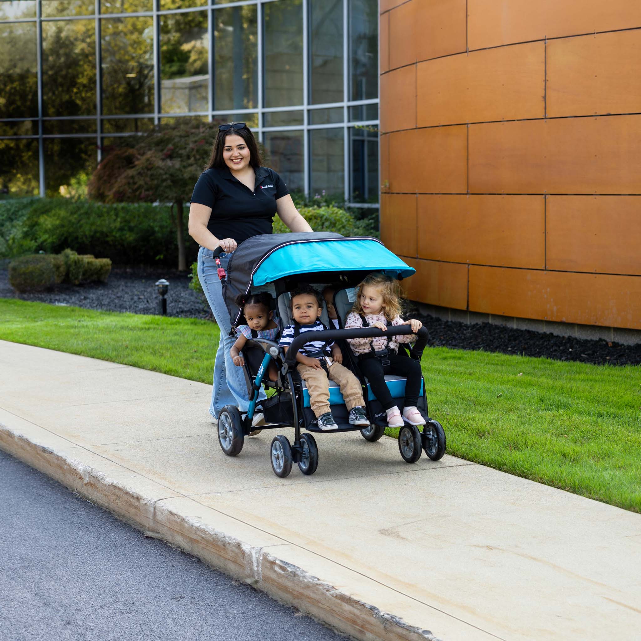Woman pushing a stroller with four children on a sidewalk, building in the background