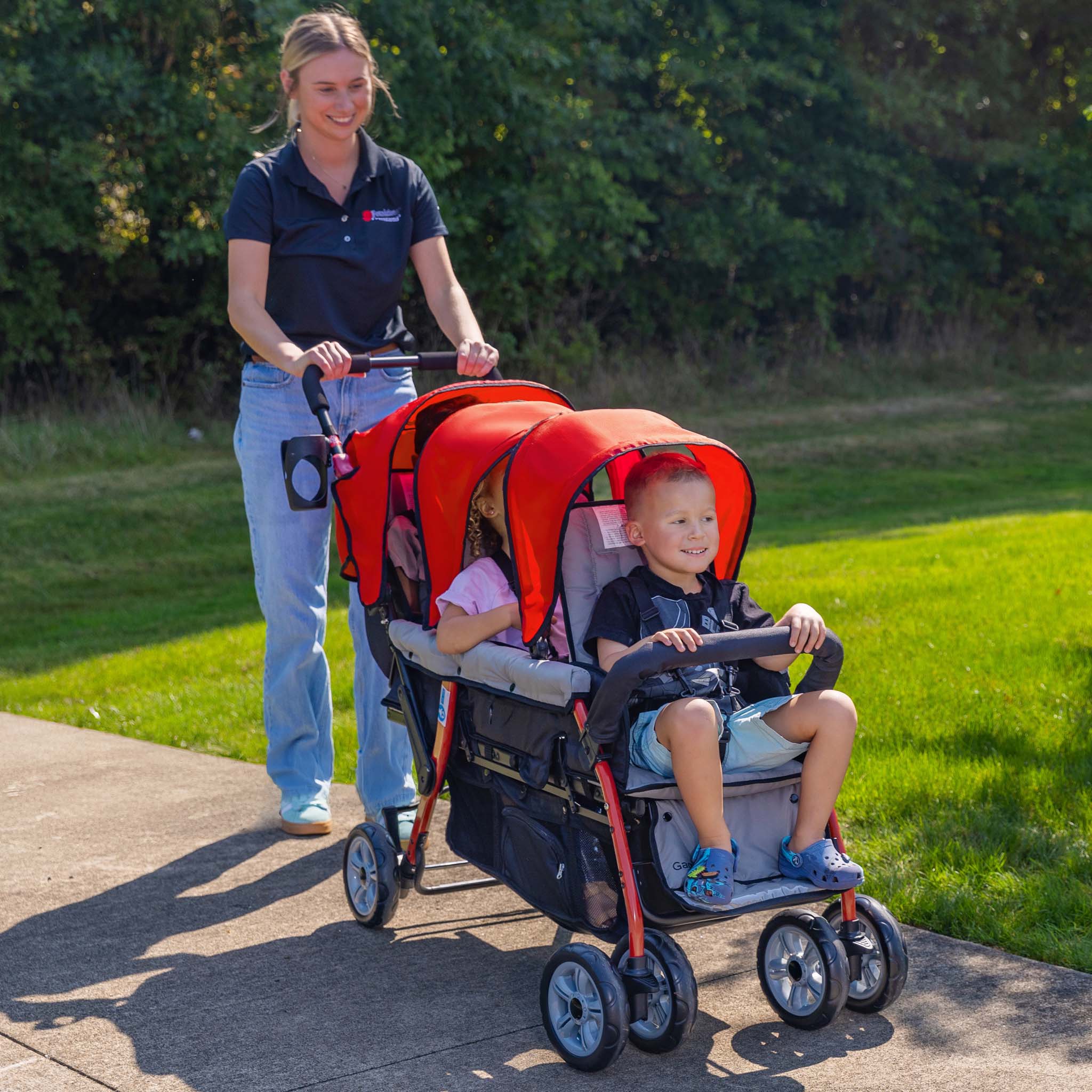 Woman pushing a red stroller with three children on a sidewalk.