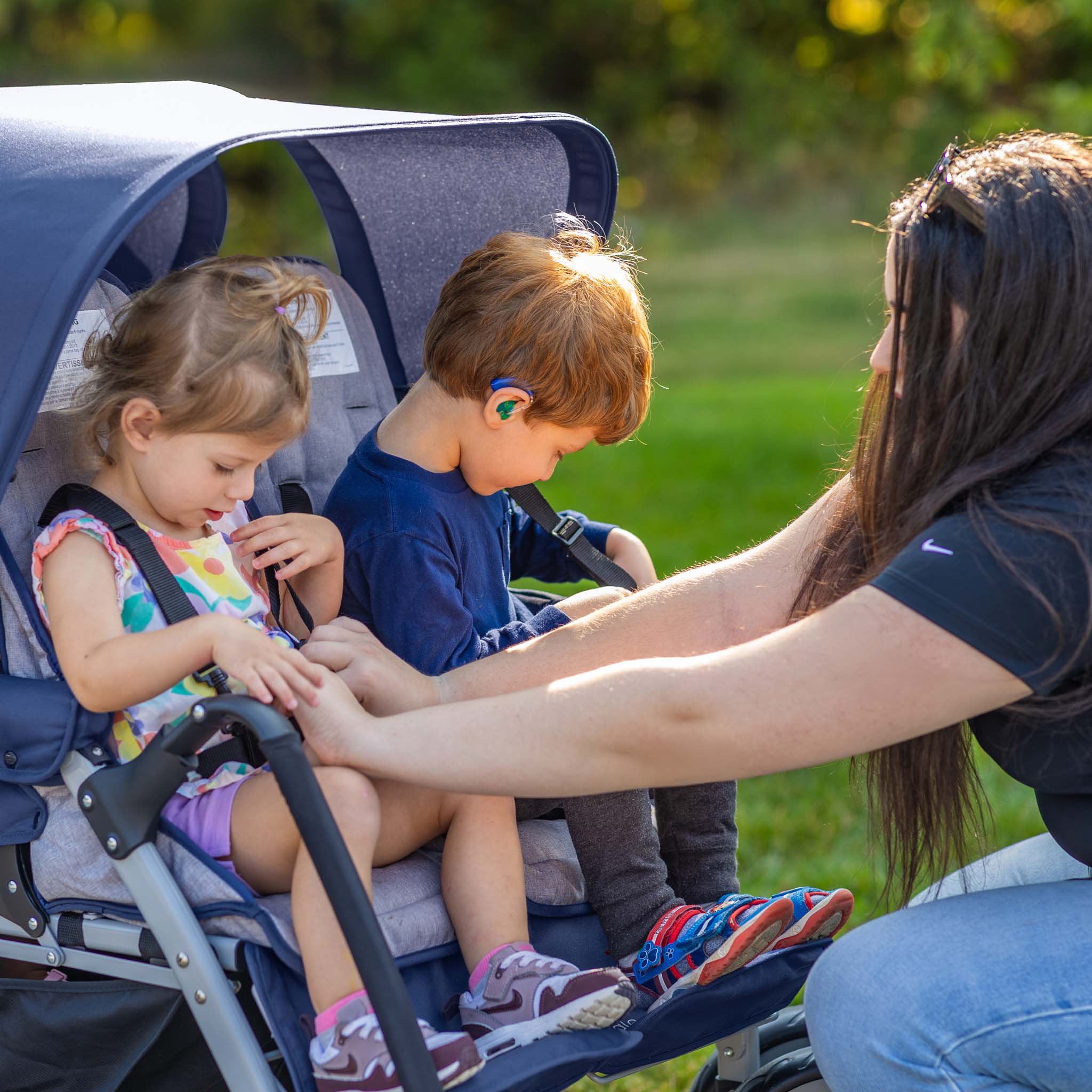 Woman adjusting a stroller with two children sitting inside, outdoors.