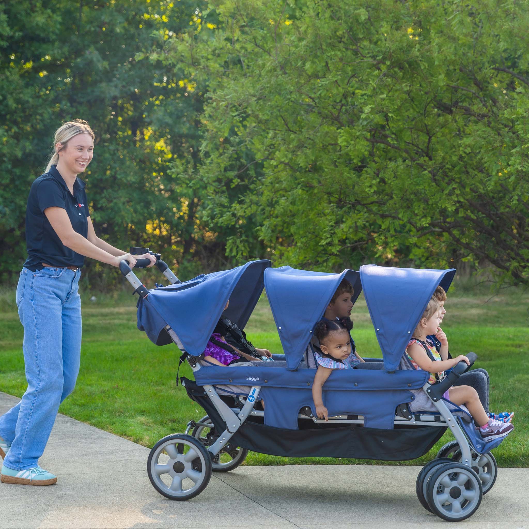 Woman pushing a blue stroller with six children in a park.