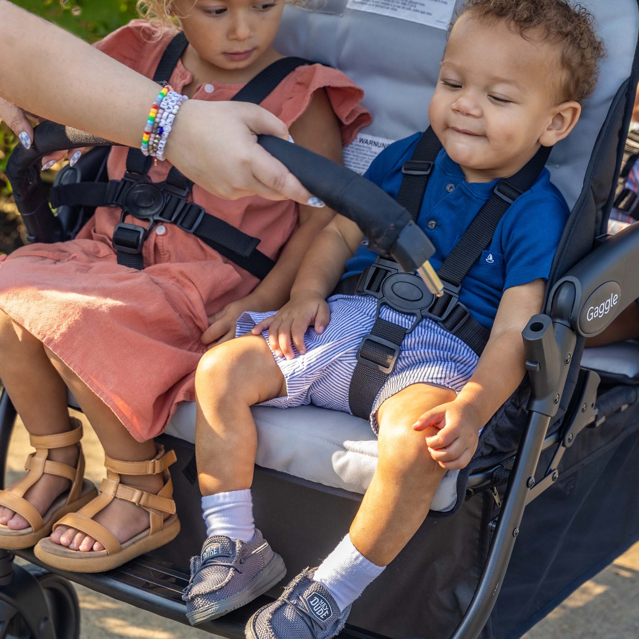 Two children in a stroller with a person adjusting arm bar outdoors.