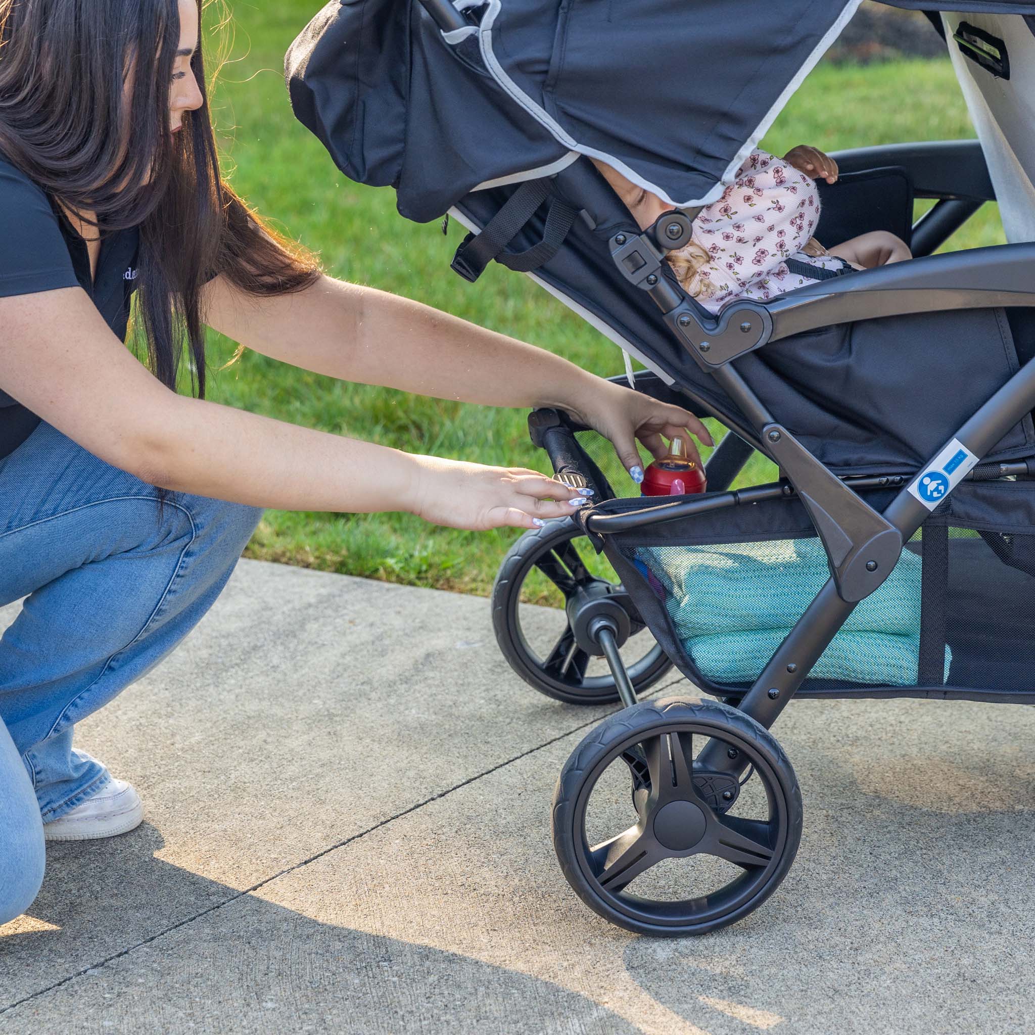 Woman adding to stroller storage basket of a 4 seat stroller on a sidewalk with grass in the background