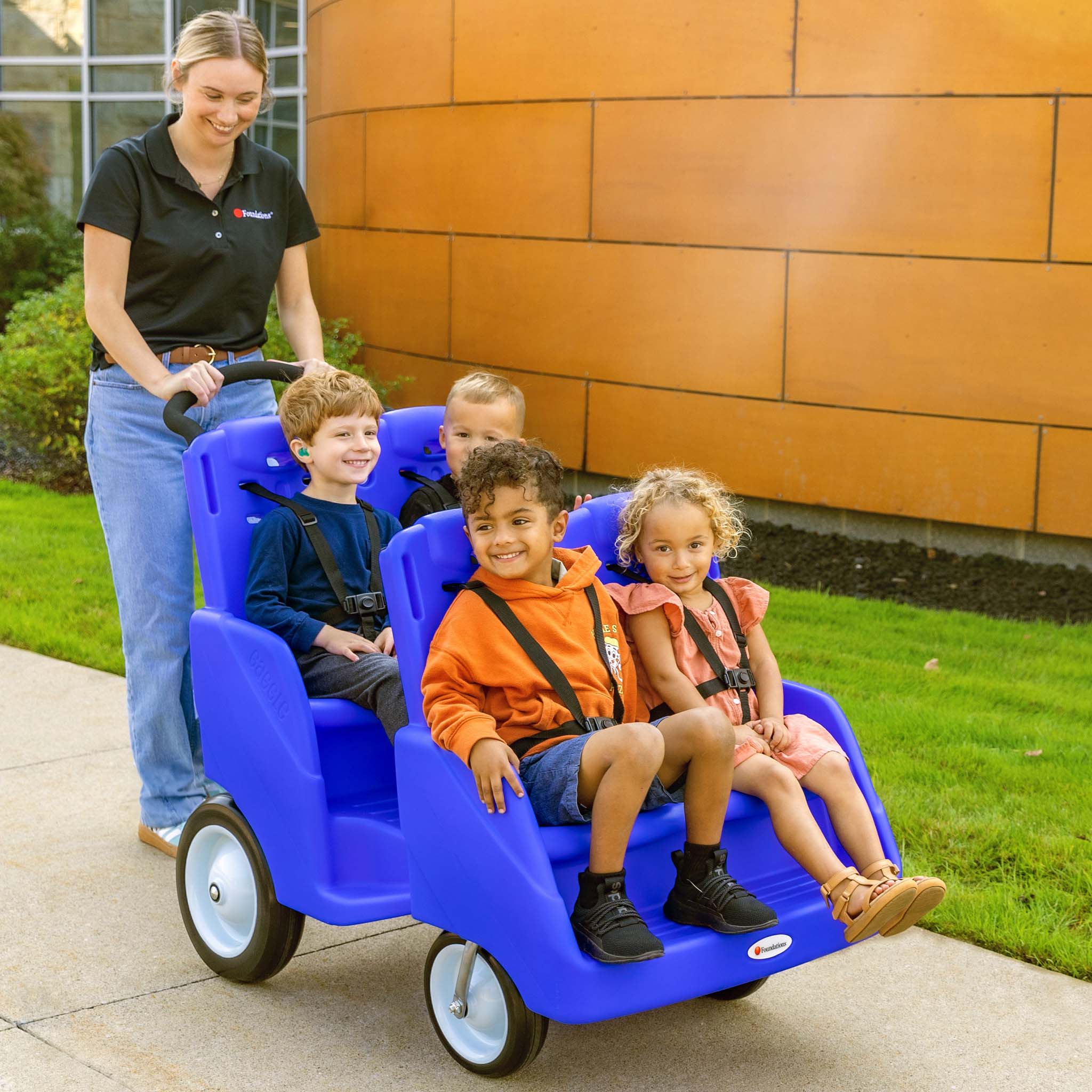 Woman pushing a blue children's wagon with four children sitting inside on a sidewalk.