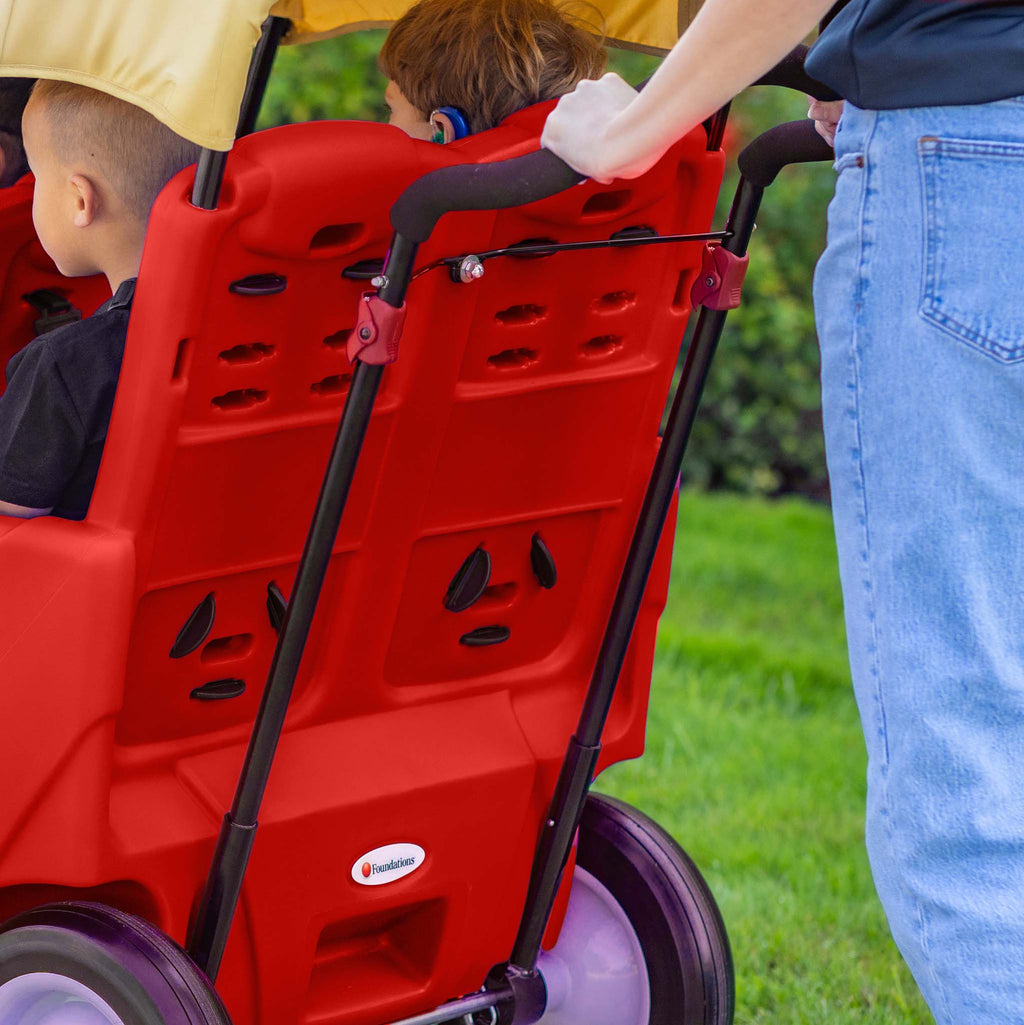 Back of four seat red wagon with children inside, being pushed by an adult on a grassy area.