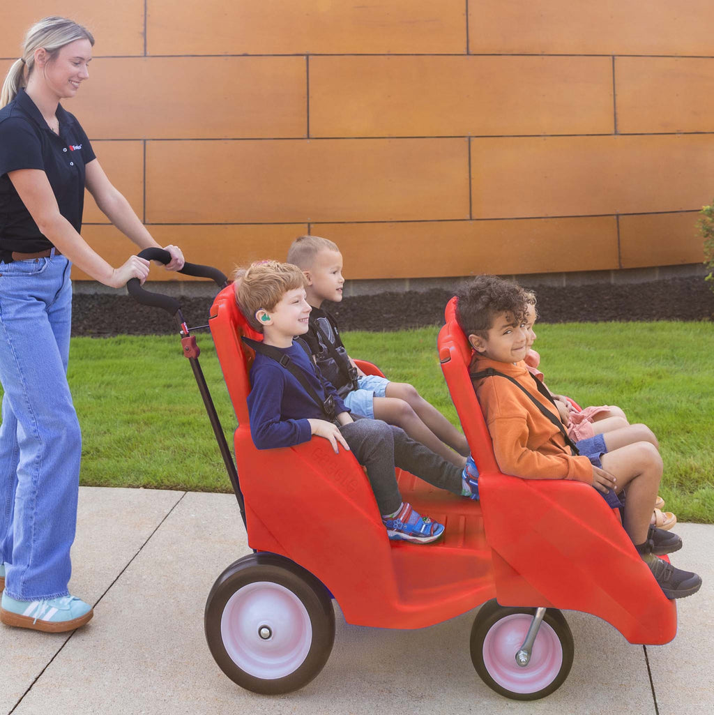 Woman pushing a red four seat children's wagon with children sitting inside on a sidewalk.