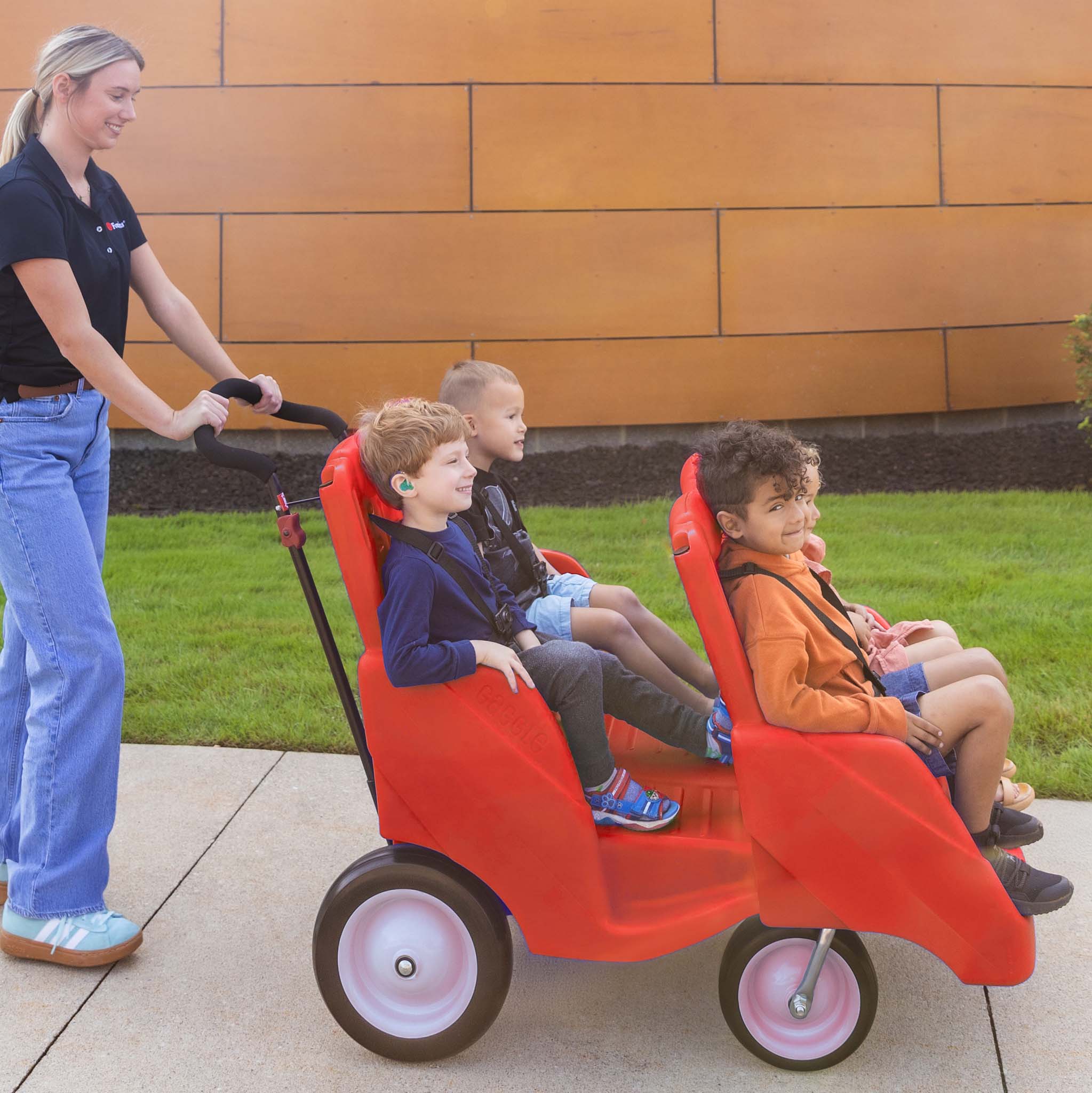 Woman pushing a red four seat children's wagon with children sitting inside on a sidewalk.